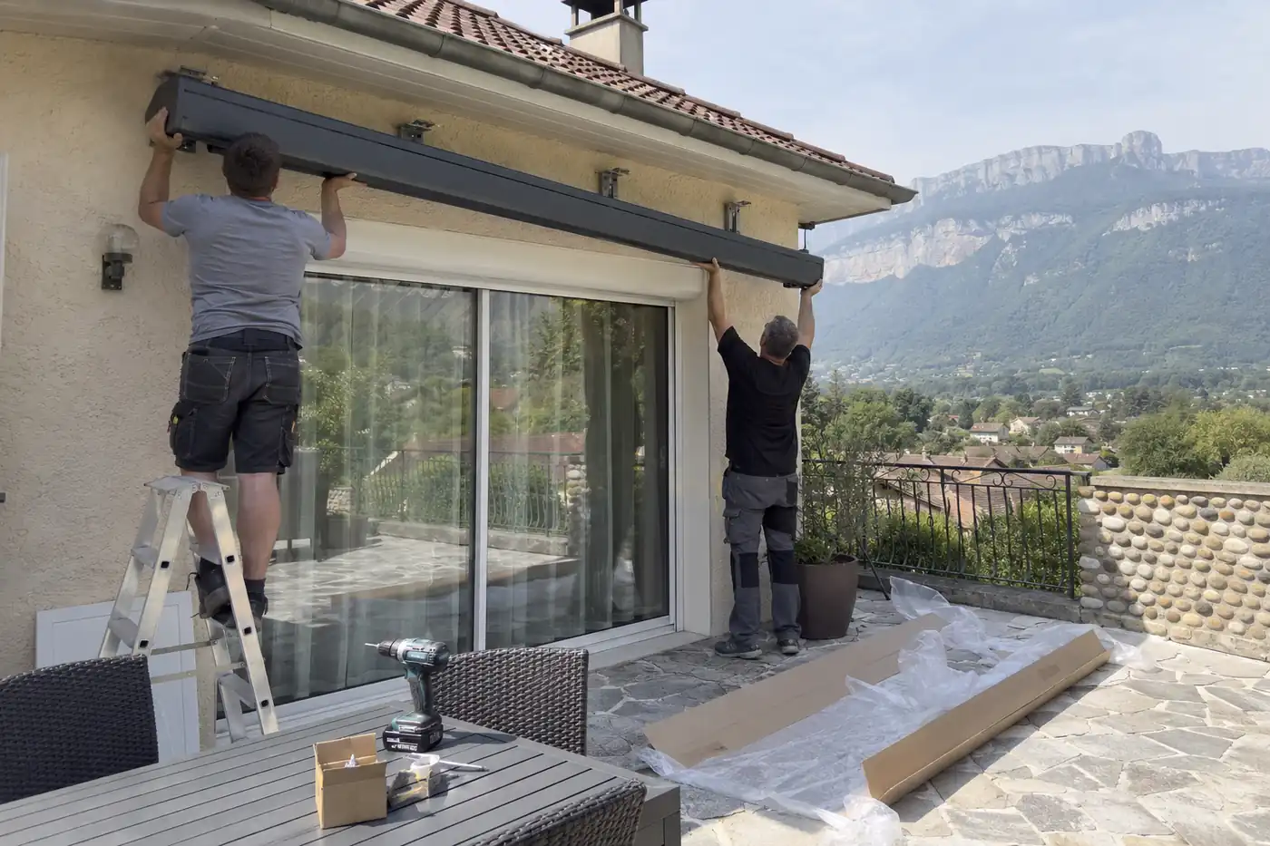 Deux poseurs levant un coffre de store banne contre la façade d’une maison, cartons et outillage sur la terrasse