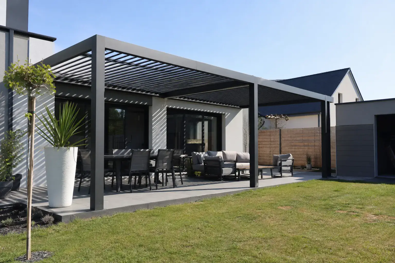 Terrasse couverte par une pergola aux lames aluminium entrouvertes, vue de fin de matinée