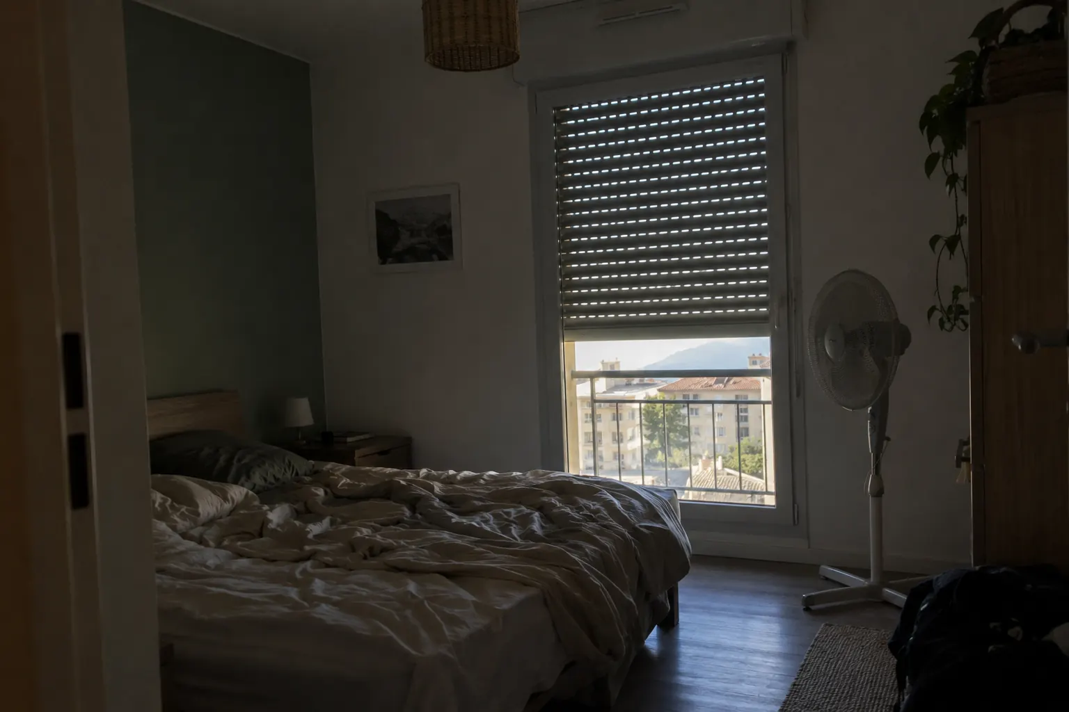 Chambre plongée dans la pénombre en été, volet roulant presque baissé devant la fenêtre, ventilateur au pied du lit