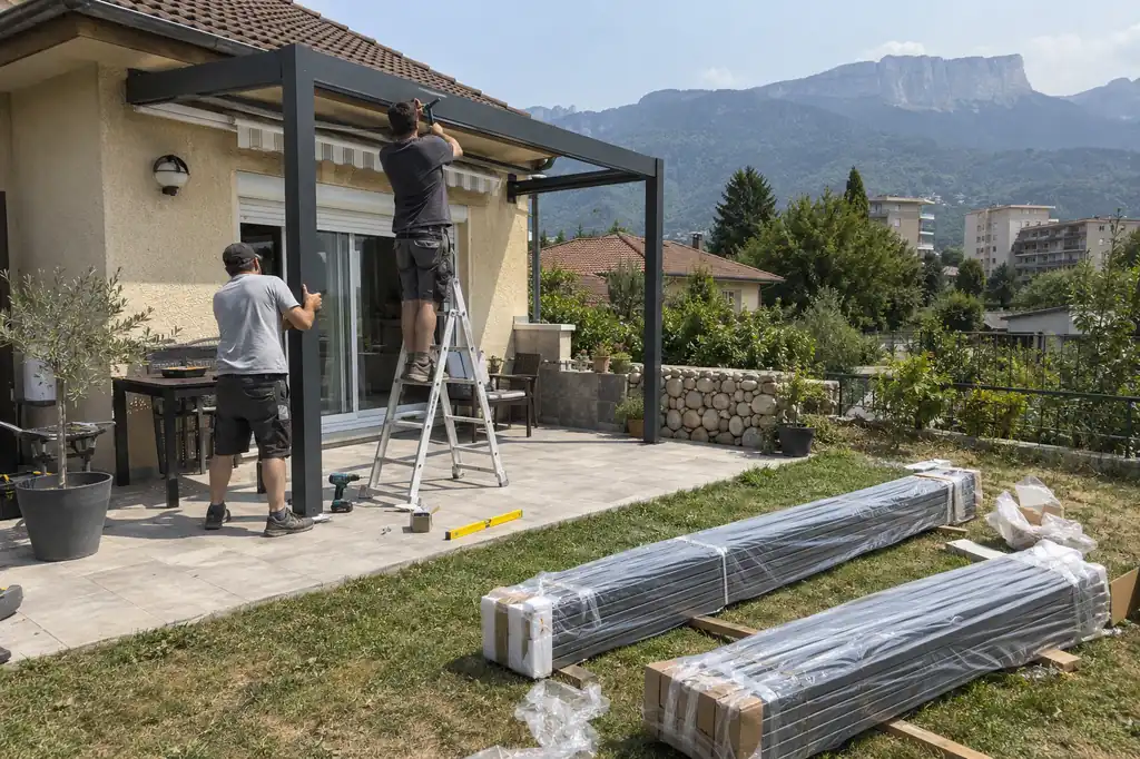 Deux monteurs fixant la structure d’une pergola contre une maison, colis de lames emballés posés sur la pelouse
