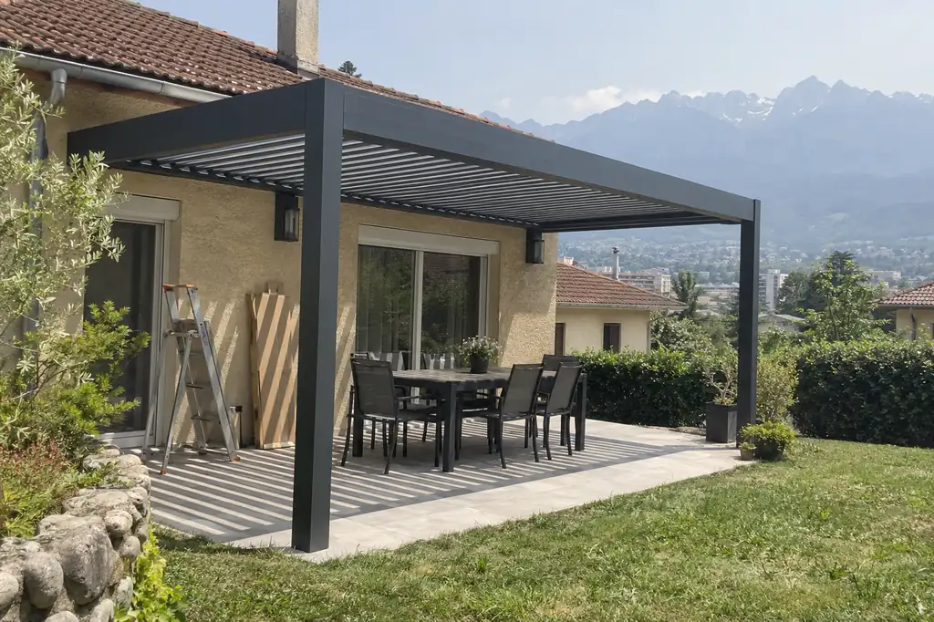 Pergola bioclimatique gris anthracite sur une terrasse de maison, table et chaises sous les lames, montagnes au fond