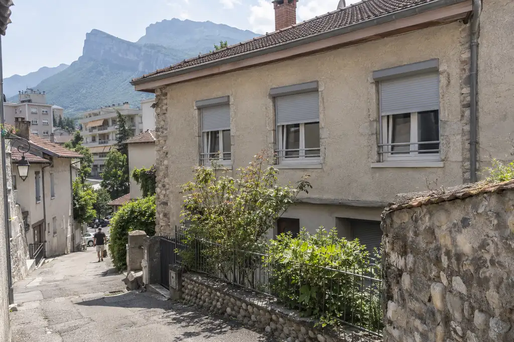 Maison de village en pierre équipée de volets roulants blancs à chaque fenêtre, ruelle en pente devant